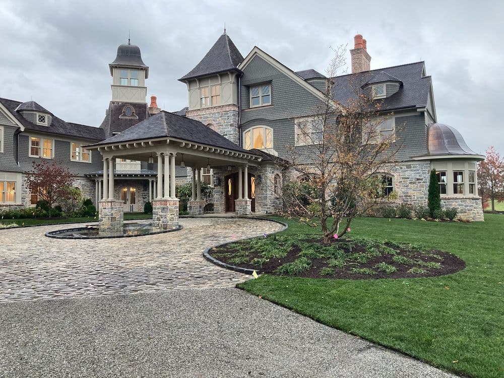 Elegant gray mansion with stone accents, landscaped driveway, and cloudy sky backdrop.
