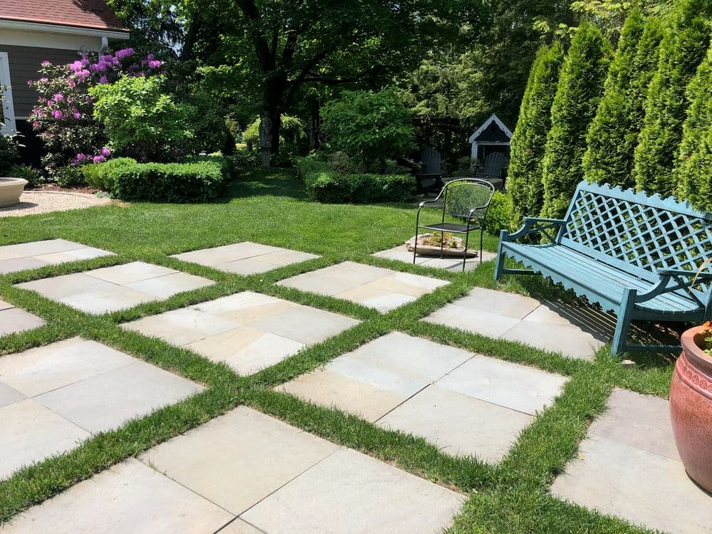 Patio with stone pavers, green grass, blue bench, and flowering bushes in a garden setting.