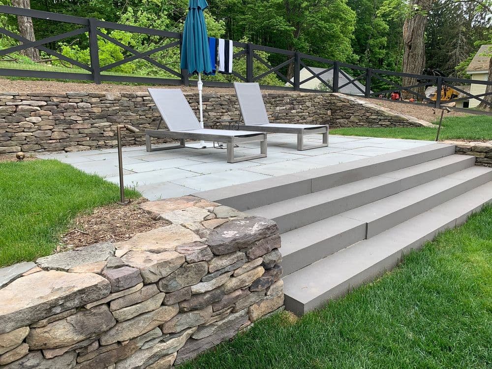Elegant outdoor patio with stone steps, lounge chairs, and a blue umbrella in a lush green setting.