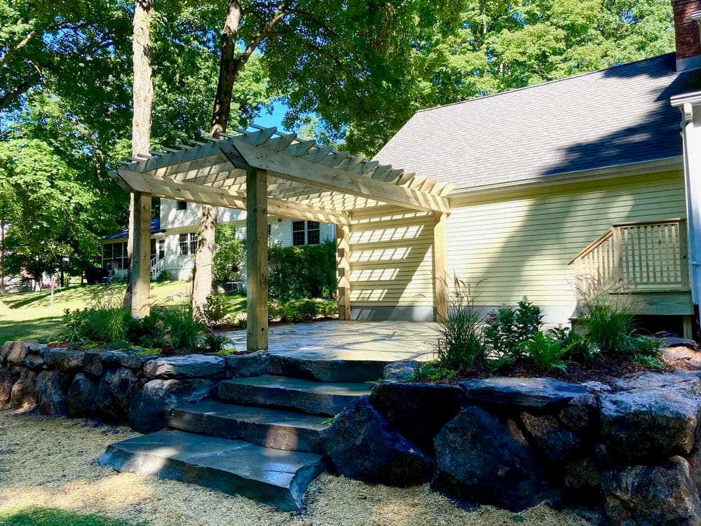 Wooden pergola over a stone patio surrounded by lush greenery and landscaped gardens.