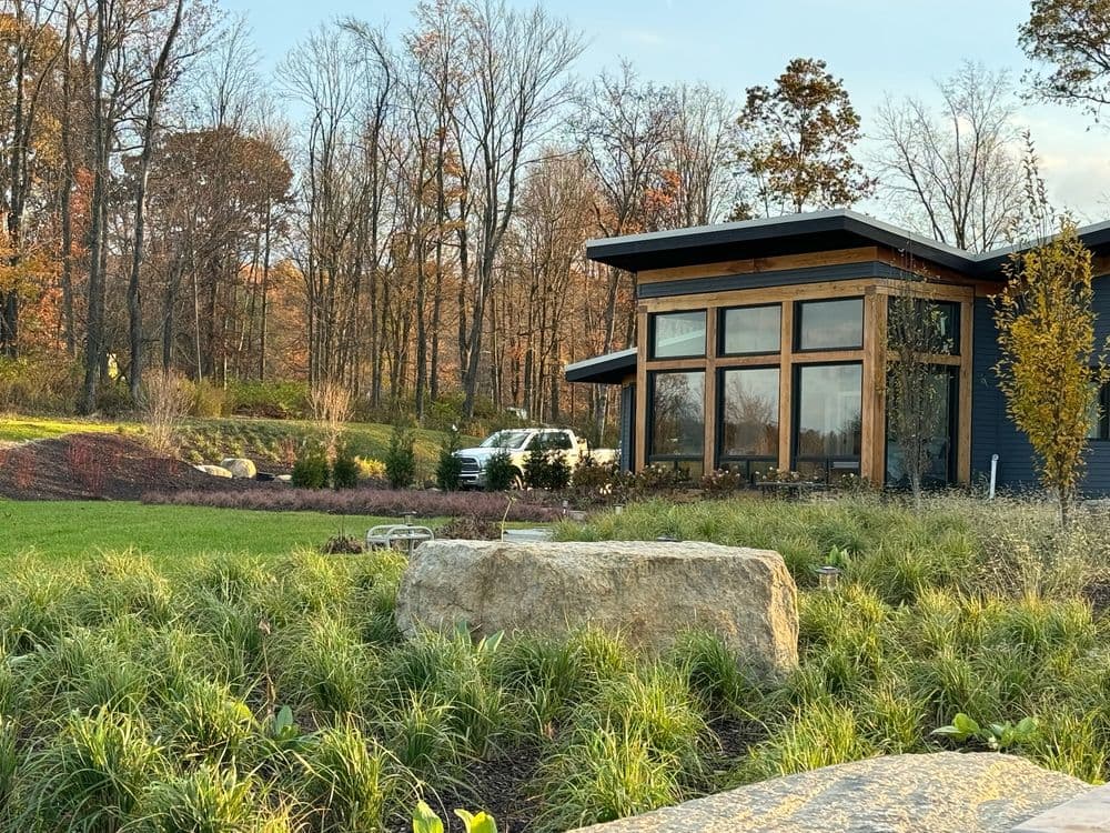 Modern house with large windows surrounded by lush landscaping and scenic autumn trees.