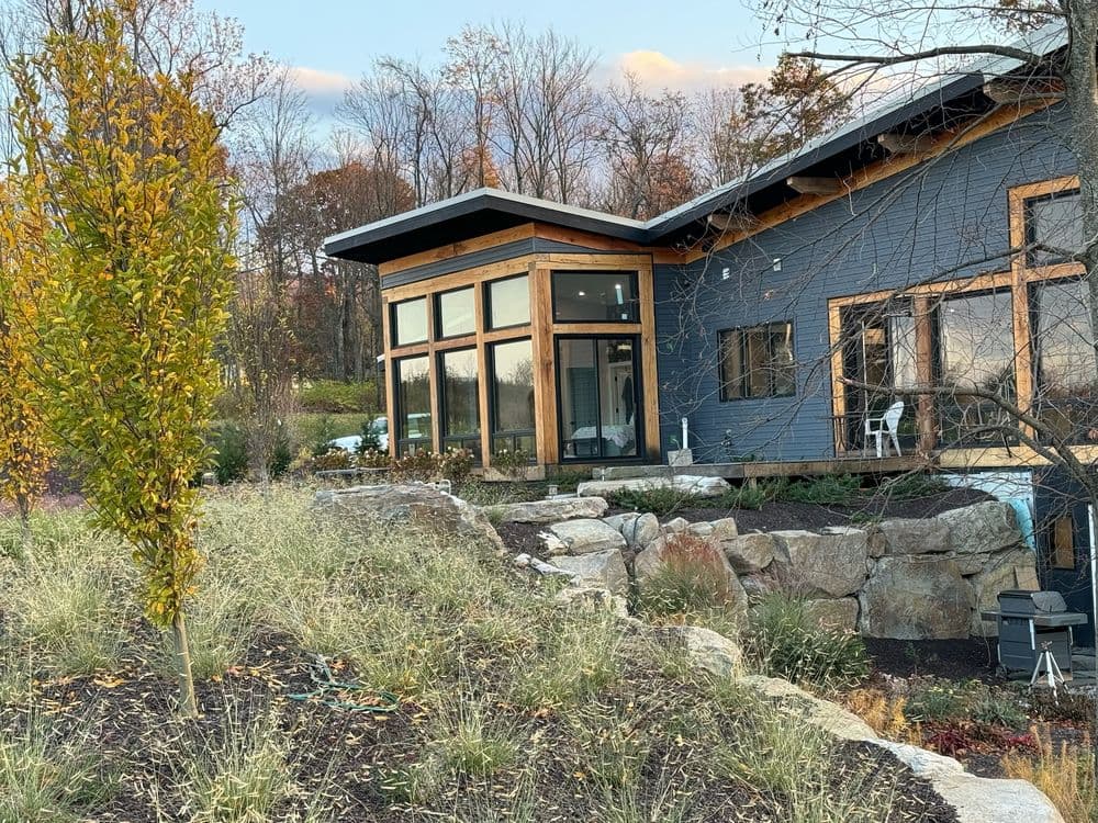 Modern house with large windows, surrounded by greenery and stones, set in a tranquil landscape.